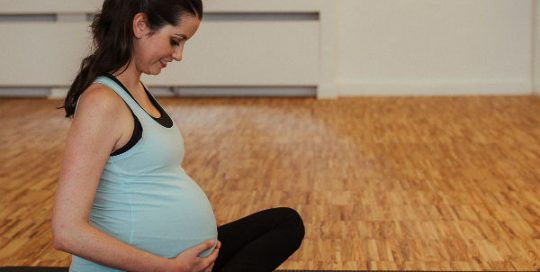 Schwangere Frau sitzt glücklich auf einer Pilates Matte und hält mit ihren Händen ihren schwangeren Bauch fest.