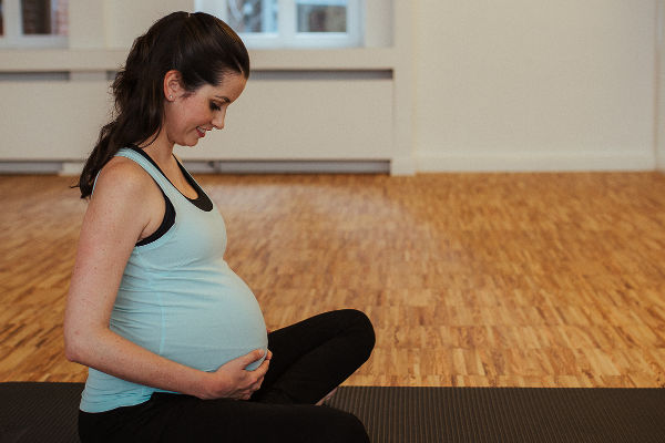 Schwangere Frau sitzt glücklich auf einer Pilates Matte und hält mit ihren Händen ihren schwangeren Bauch fest.